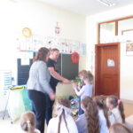 Przedstawiciele firmy recyklingowej wręczają dzieciom nagrody za poprawne odpowiedzi podczas spotkania o segregacji śmieci.