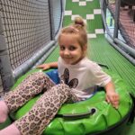 Dziewczynka w białej koszulce i spodniach w panterkę siedzi uśmiechnięta w zielonym pontonie na zjeżdżalni w sali zabaw.