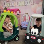 dwóch chłopców jedzie małymi plastikowymi samochodami – jeden zielonym z dinozaurem, drugi czarnym z uśmiechniętą buzią na przodzie.