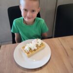 Na zdjęciu widać chłopca siedzącego przy stole. Ma krótkie włosy i zieloną koszulkę, patrzy w stronę aparatu z lekkim uśmiechem. Przed nim, na białym talerzu, leży gofr udekorowany bitą śmietaną, kawałkami owoców, kolorowymi posypkami oraz pianką marshmallow