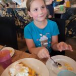 Dziewczynka w niebieskiej koszulce siedzi przy stole, obok talerz z gofrem udekorowanym bitą śmietaną, owocami i posypką.