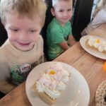 dzieci przy stole, przed nimi talerze z udekorowanymi goframi. Gofry pokryte są bitą śmietaną, owocami, piankami i kolorowymi posypkami.