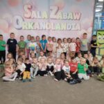 Grupa przedszkolaków pozuje wspólnie na tle napisu „Sala Zabaw Orkanolandia”. Dzieci uśmiechają się i pozują w dwóch rzędach – część siedzi, a część stoi.