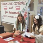 Dwie uczennice ubrane w stroje stylizowane na renesansowe siedzą przy okrągłym stole przykrytym czerwonym obrusem. Na stole stoją dwie złote świeczniki, kilka książek i podręczników. W tle widać tablicę z napisem „Narodowe Czytanie 2025 Jan Kochanowski – Treny, Fraszki, Pieśni”