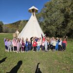 Grupa przedszkolaków pozuje do wspólnego zdjęcia na tle dużego tipi. Dzieci stoją na trawie, machają i uśmiechają się do obiektywu. W tle widać jesienne drzewa i błękitne niebo.