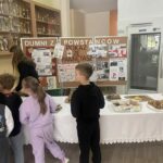 Grupa dzieci ogląda i kupuje ciasta na kiermaszu charytatywnym zorganizowanym w szkole. W tle widoczne są gabloty z pucharami sportowymi oraz tablice z pracami o Powstaniu Warszawskim.