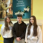 Troje uczniów stoi na tle szkolnej tablicy z wizerunkiem św. Józefa oraz napisem o jego cechach. Za nimi widać żółte szafki szkolne. Uczniowie patrzą w obiektyw i uśmiechają się delikatnie. Zdjęcie przedstawia przedstawicieli nowo wybranego Samorządu Uczniowskiego.