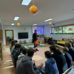 Na zdjęciu widać grupę uczniów siedzących na ławkach i uważnie słuchających instruktora ubranego w strój ratownika medycznego. W sali, ozdobionej żółtymi papierowymi kulami i jesiennymi dekoracjami, prowadzone są warsztaty z pierwszej pomocy. Na podłodze leżą fantomy, na których uczestnicy będą ćwiczyć resuscytację. W tle widoczny jest ekran z prezentacją zatytułowaną „Bezpieczni na stoku – warsztaty z pierwszej pomocy