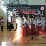 Dziewczynki w czerwonych spódnicach i białych bluzkach śpiewają przy mikrofonach podczas akademii.