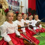 Pięć młodych dziewczynek w białych bluzkach z haftowanymi kołnierzami i czerwonych spódnicach siedzi w rzędzie na scenie, trzymając skrzypce. Każda ma we włosach czerwoną ozdobę. Za nimi znajduje się dekoracja w barwach biało-czerwonych oraz sylwetka powstańca, tworząca patriotyczne tło