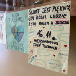 Uczniowski plakat na Dzień Tolerancji z hasłami o zgodzie, empatii i życzliwości, zawieszony pod szkolnymi pucharami.