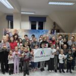 Grupowe zdjęcie dzieci i dorosłych w sali warsztatowej. Uczestnicy stoją w kilku rzędach, uśmiechnięci, trzymają w dłoniach własnoręcznie wykonane, kolorowe ozdoby świąteczne. Na środku grupa dzieci prezentuje baner z napisem „Małopolska”. W tle widać dekoracje zimowe, tablice oraz jasne oświetlenie sali, tworzące ciepłą, wspólnotową atmosferę twórczych warsztatów.