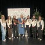 Grupa uczennic oraz opiekunka stoją na scenie podczas V Gali Wolontariatu 2025; w tle dekoracja z logo wydarzenia i choinki.