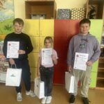 Na zdjęciu stoi trójka uczniów w sali szkolnej na tle kolorowych szafek. Dzieci trzymają dyplomy i białe torby z nagrodami. Od lewej: Karol Kowalczyk (kl. V), Anna Kowalczyk (kl. II) oraz Jakub Mikołajczyk (kl. V) – laureaci konkursu plastycznego „Reymontowska wieś w obrazach” organizowanego przez Gminną Bibliotekę Publiczną.