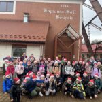 Duża grupa dzieci i opiekunów w kolorowych zimowych kurtkach i czerwonych czapkach mikołajowych pozuje do wspólnego zdjęcia przed wejściem do Kopalni Soli w Bochni. W tle widać budynek z napisem ‘Kopalnia Soli Bochnia 1248’ oraz fragment konstrukcji szybu. Uczestnicy stoją i kucają w kilku rzędach, uśmiechnięci, gotowi do rozpoczęcia świątecznej przygody.