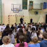 Uczniowie zgromadzeni na sali gimnastycznej siedzą razem i słuchają prowadzących spotkanie policjantów. Wspólna obecność i uważność tworzą atmosferę skupienia, rozmowy.