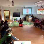 Uczniowie klas czwartych i piątych siedzą w kręgu w sali szkolnej podczas warsztatów z pierwszej pomocy; prowadząca klęczy na środku i instruuje dzieci. W tle widoczny ekran z prezentacją oraz wyposażenie sali.