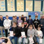 Grupowe zdjęcie uczniów i nauczycieli w sali szkolnej. Uczniowie stoją i klęczą w dwóch rzędach, trzymają dyplomy i mają zawieszone na szyi medale. W tle widoczne plansze edukacyjne z matematyki oraz baner instytucji. Zdjęcie wykonane podczas uroczystości wręczenia nagród.