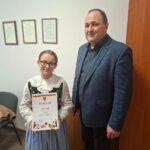 Uczennica w tradycyjnym stroju regionalnym stoi obok dorosłego mężczyzny w marynarce. Dziewczynka trzyma dyplom za zajęcie II miejsca w konkursie „Ekologiczny Stroik Bożonarodzeniowy”. Oboje patrzą w stronę aparatu.