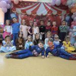 Grupa przedszkolaków w kolorowych strojach karnawałowych pozuje do wspólnego zdjęcia na tle dekoracyjnej ścianki z balonami.