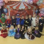 Grupa przedszkolaków w kolorowych strojach karnawałowych pozuje do wspólnego zdjęcia na tle dekoracyjnej ścianki z balonami.