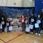 Grupowe zdjęcie uczniów i nauczycieli w sali gimnastycznej podczas szkolnej uroczystości. Dzieci trzymają dyplomy i torby z upominkami, stojąc na tle świątecznej dekoracji z szopką, choinkami i zimową scenerią.