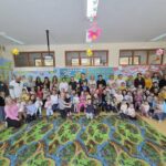 Na zdjęciu widać dużą grupę dzieci z oddziałów przedszkolnych oraz ich rodziców i nauczycieli, którzy wspólnie pozują do pamiątkowego zdjęcia w kolorowo udekorowanej sali przedszkolnej. Dzieci trzymają w rękach własnoręcznie wykonane ozdoby wielkanocne. Sala jest ozdobiona wiosennymi dekoracjami, takimi jak kwiaty i motyle zwisające z sufitu oraz kolorowe prace plastyczne na ścianach. Wszyscy siedzą i stoją na dużym, kolorowym dywanie z motywem rzeki i lasu. Atmosfera zdjęcia jest radosna i rodzinna, a uczestnicy uśmiechają się do wspólnej fotografii.