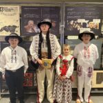 Czworo uczniów w tradycyjnych strojach góralskich stoi przed wystawą edukacyjną; jeden z chłopców trzyma książkę „Zagórzańska cytanka”.