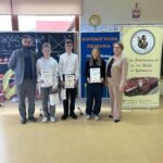 Laureaci konkursu „Matematyczne Zmagania” z dyplomami i nagrodami stoją wraz z organizatorami na tle tablicy i banerów szkolnych.