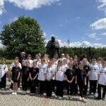 Grupa dzieci i opiekunów z zespołu regionalnego „Śwarni Lubomiyrzanie” stoi na placu przed pomnikiem św. Jana Pawła II w Ludźmierzu. Uczestnicy ubrani są w białe i czarne koszulki z nazwą zespołu. W tle widoczny jest rozległy teren sanktuarium z zielenią, drzewami oraz błękitnym niebem z białymi chmurami. Zdjęcie wykonano podczas Warsztatów Zagórzańskich poświęconych poznawaniu kultury, tradycji i dziedzictwa Zagórzańszczyzny.