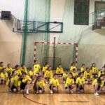 Uczennice prezentują występ taneczny na sali gimnastycznej. Dzieci w żółtych koszulkach poruszają się w zorganizowanym układzie choreograficznym.