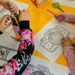 Dzieci siedzą przy żółtym stoliku i kolorują obrazki przedstawiające aparat fotograficzny ozdobiony sercami. Na stole znajdują się kredki, flamastry i nożyczki. Uczestnicy warsztatów z zaangażowaniem tworzą prace związane z tematem „Aparat ważnych chwil”, rozwijając wyobraźnię i rozmawiając o ważnych dla nich momentach spędzanych z bliskimi.