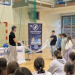 Uczniowie zgromadzeni w sali gimnastycznej obserwują pokaz naukowy Grupy „Zaelektryzowani”. Prowadzący wraz z kilkorgiem uczniów prezentuje doświadczenie z wykorzystaniem długiej foliowej tuby.