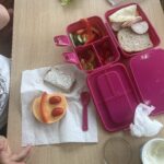 Na drewnianym stole znajdują się produkty przygotowane przez uczniów klasy trzeciej podczas zajęć o zdrowym odżywianiu. W różowej śniadaniówce widać pokrojone ogórki, czerwoną paprykę, pomidorki, pieczywo, ser i wędlinę. Obok, na papierowym ręczniku, leży kromka chleba z serkiem oraz kolorowa kompozycja z sera, papryki i pomidorków, przypominająca uśmiechniętą buzię. Zdjęcie przedstawia kreatywne przygotowywanie zdrowego drugiego śniadania.