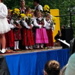 Dziewczynki w strojach ludowych trzymają żółte kwiaty podczas występu na scenie Gorczańskiego Spotkania z Janem Pawłem II. W tle widać nauczycielki w regionalnych strojach.