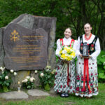 Dwie dziewczyny w tradycyjnych strojach ludowych stoją przy pomniku Jana Pawła II w Lubomierzu-Rzekach. Jedna trzyma bukiet żółtych kwiatów, druga – latarenkę.