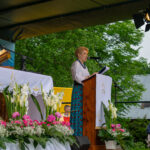 Na tle góralskiej sceny i zieleni brzóz kobieta wygłasza przemówienie podczas Gorczańskiego Spotkania z Janem Pawłem II.