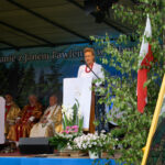 Kobieta śpiewa psalm z mównicy ozdobionej białym obrusem i kwiatami podczas Gorczańskiego Spotkania z Janem Pawłem II w Lubomierzu-Rzekach. W tle siedzą kapłani w złotych i czerwonych ornatach.