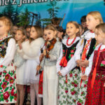 Grupa dziewczynek w tradycyjnych strojach ludowych oraz eleganckich białych ubraniach występuje na scenie podczas Gorczańskiego Spotkania z Janem Pawłem II. Jedna z dziewczynek trzyma skrzypce.