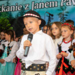 Chłopiec w góralskim stroju recytuje do mikrofonu podczas Gorczańskiego Spotkania z Janem Pawłem II. Za nim stoją dziewczynki w ludowych strojach i białych ubraniach.