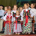 Grupa dziewczynek w tradycyjnych strojach ludowych oraz eleganckich białych ubraniach występuje na scenie podczas Gorczańskiego Spotkania z Janem Pawłem II.