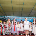 Dzieci w strojach regionalnych i białych uroczystych strojach stoją na scenie podczas Gorczańskiego Spotkania z Janem Pawłem II w Lubomierzu-Rzekach. Część trzyma kwiaty, inne śpiewają do mikrofonów.