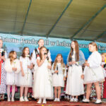 Dzieci w strojach regionalnych i białych uroczystych strojach stoją na scenie podczas Gorczańskiego Spotkania z Janem Pawłem II w Lubomierzu-Rzekach. Część trzyma kwiaty, inne śpiewają do mikrofonów.