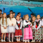 Grupa dzieci w strojach regionalnych i eleganckich ubraniach występuje na scenie podczas Gorczańskiego Spotkania z Janem Pawłem II w Lubomierzu-Rzekach.