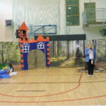 Kobieta przemawia na tle dekoracji lasu i zamku podczas przedstawienia „Szewczyk Dratewka” na sali gimnastycznej.