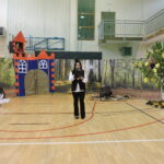 Kobieta- narrator czyta podczas występu „Szewczyk Dratewka” na tle dekoracji lasu, zamku i drzewa z pszczołami.