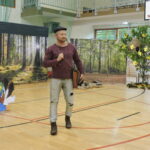 Mężczyzna w stroju Szewczyka Dratewki stoi na sali gimnastycznej, w tle dekoracje lasu, zamek i drzewo z pszczołami.