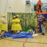 Szewczyk Dratewka stoi przy dekoracyjnym zamku, naprzeciwko grupy dorosłych przebranych za kaczuszki, w tle księżniczka spogląda z wieży.