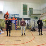 Trzech dorosłych mężczyzn odgrywa scenę na sali gimnastycznej – w tle zamek z wieżą, z której wygląda księżniczka, oraz dekoracje lasu i drzewa z papierowymi pszczołami.