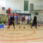 Trzech dorosłych mężczyzn odgrywa scenę na sali gimnastycznej – w tle zamek z wieżą, z której wygląda księżniczka, oraz dekoracje lasu i drzewa z papierowymi pszczołami.